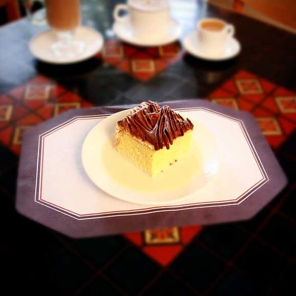 A slice of cake with chocolate frosting on a white plate, with coffee cups in the background.