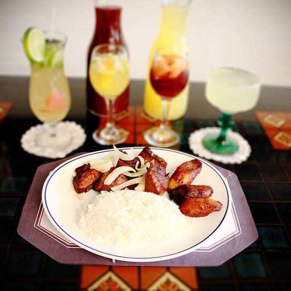 Cuban dish with rice, fried pork chunks, plantains, and onions, surrounded by colorful drinks.