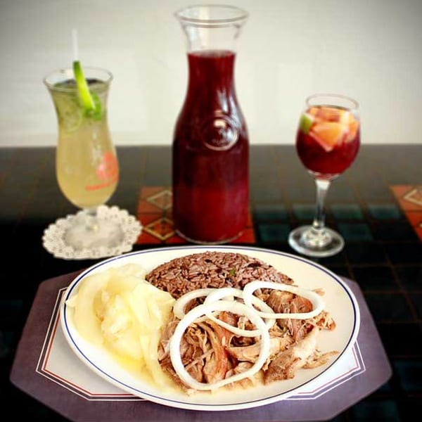 Cuban roasted pork with onions, rice, and plantains, served with a pitcher and glasses of red and yellow drinks.