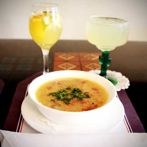 A bowl of Cuban soup with parsley, alongside two glasses of refreshing drinks on a table.