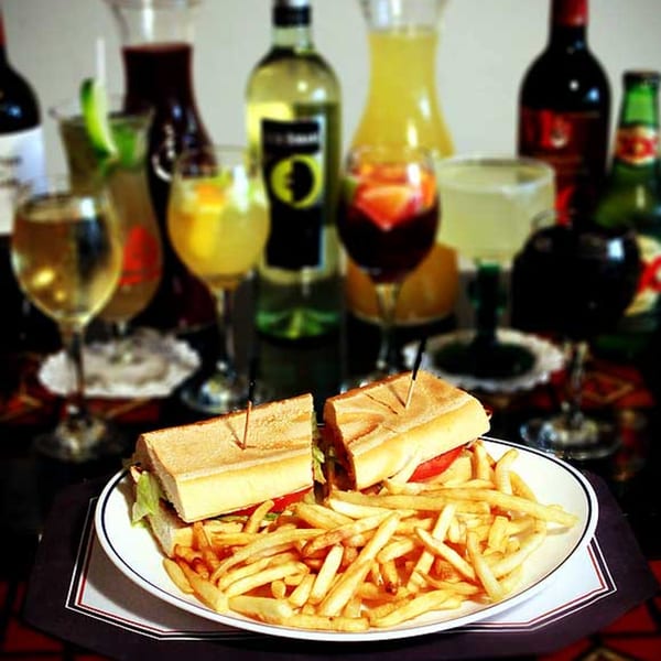 A Cuban sandwich with fries on a white plate, surrounded by various drinks and bottles.