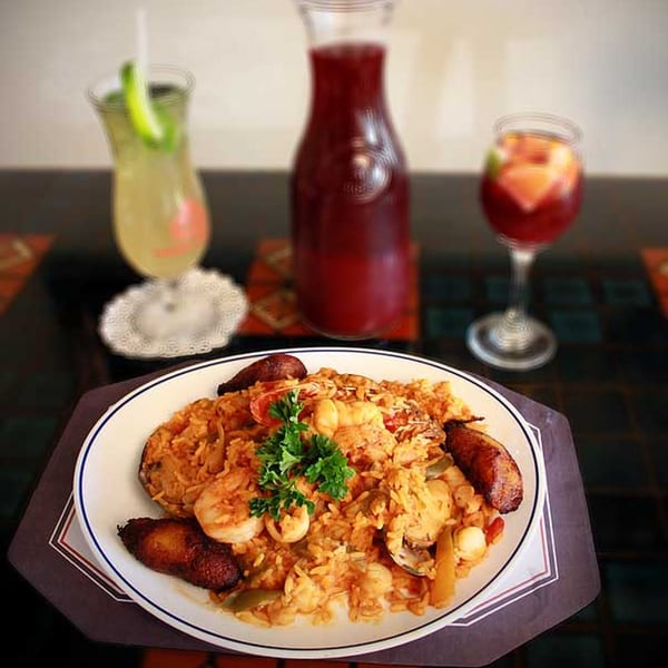 Cuban seafood paella with plantains, garnished with parsley, and two drinks on a dark table.